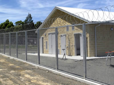 358 anti-climb fencing system surrounds a yellow factory with razor wires on the top of it.